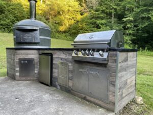 Cabin in the Woods door on a Forno Nardona wood-fired Napoli brick oven in green with white stripes on a rustic wood outdoor kitchen with a black poured concrete countertop featuring a Lion grill, refrigerator and doors/drawers.