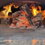 Red hot wood coals ready to be scattered in a wood fired oven by Forno Nardona with flames in the background.