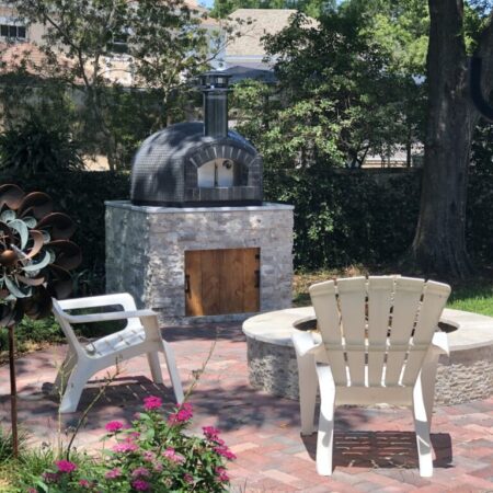 32" Nardona Rustico in grey tile sits atop a custom base also by Forno Nardona with woods in the background and a fire pit with chairs in the foreground.
