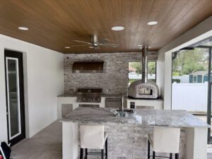 A rustic outdoor pizza kitchen under a covered space with a wood ceiling with canned lights and a ceiling fan. The kitchen features a Forno Nardona Rustico pizza oven with a washed red brick face and a Lion Premium grill with a large custom island with seating in the foreground.